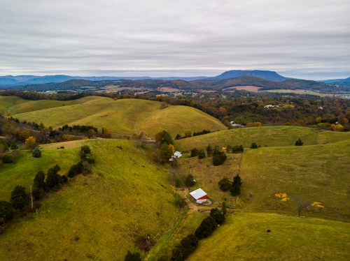 Aerial view of rolling green hills with a red barn, under a cloudy sky and distant mountains.
