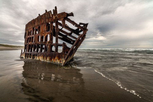 A rusted shipwreck partially submerged in a sandy beach, with waves lapping at its weathered hull under a cloudy sky.