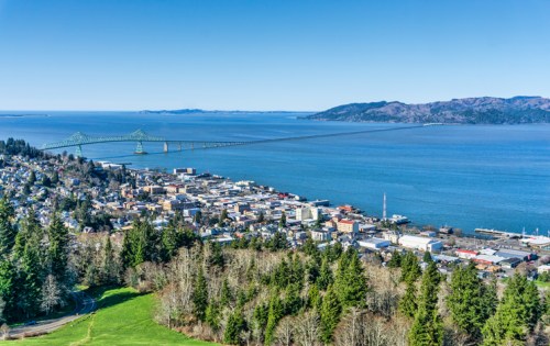 Aerial view of a coastal town with a bridge spanning the water, surrounded by hills and greenery under a clear blue sky.