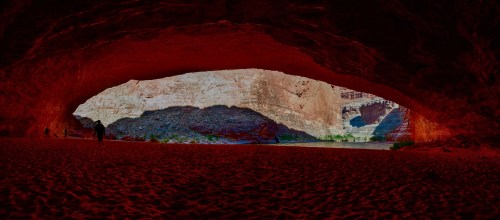 A panoramic view of a large cave opening, revealing colorful rock formations and a sandy floor.