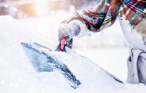 A person scrapes snow off a car window while wearing a colorful scarf and gloves in a snowy environment.