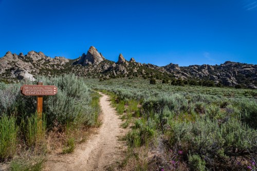 A dirt path leads through sagebrush towards rocky mountains under a clear blue sky, with a signpost nearby.