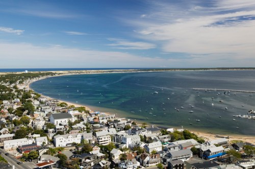 Aerial view of a coastal town with boats in the water and a sandy beach under a blue sky with scattered clouds.