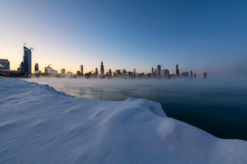 A panoramic view of a snowy Chicago skyline at sunrise, with mist rising from the icy lake.