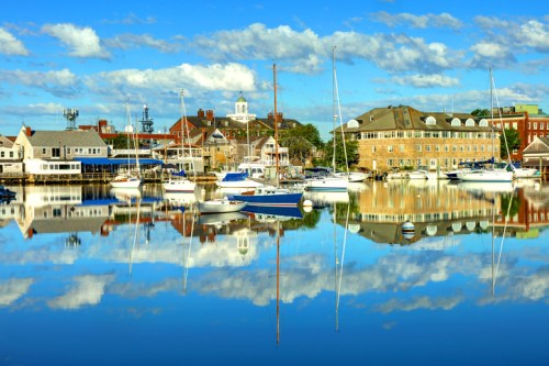 A serene harbor scene with boats reflecting on calm water, surrounded by buildings and a blue sky with fluffy clouds.
