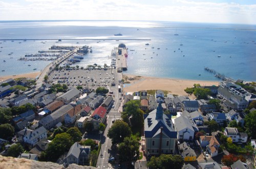 Aerial view of a coastal town with a marina, beach, and boats on the water under a clear blue sky.
