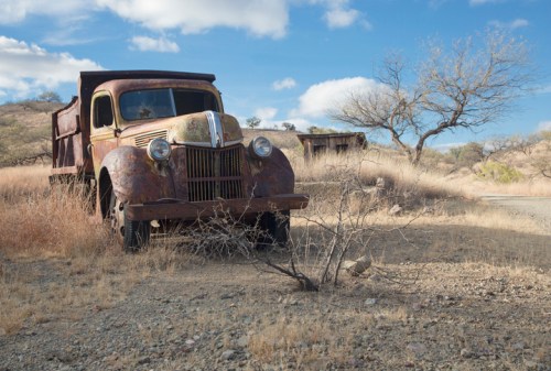 An old, rusty truck sits in a dry, grassy landscape under a blue sky with scattered clouds and a bare tree nearby.