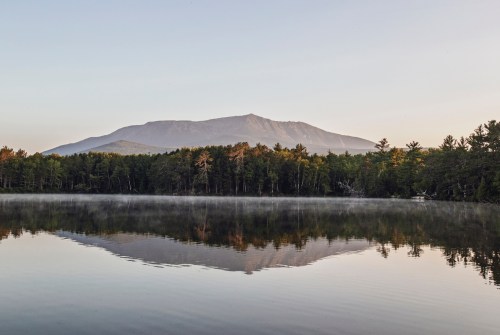 A serene lake reflects a mountain and trees at dawn, with mist rising from the water's surface.