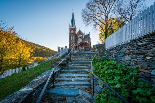 A stone pathway leads up to a historic church surrounded by trees and a white picket fence under a clear blue sky.