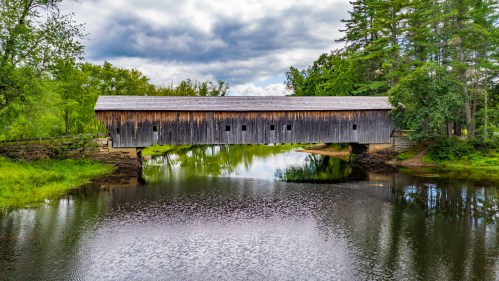 A wooden covered bridge spans a calm river, surrounded by lush greenery and a cloudy sky.