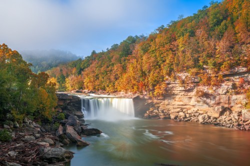 A serene waterfall surrounded by vibrant autumn foliage and misty mountains under a clear blue sky.