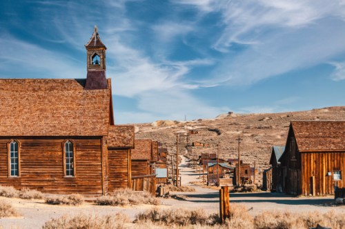 A deserted street in a ghost town, featuring wooden buildings and a church under a blue sky with wispy clouds.