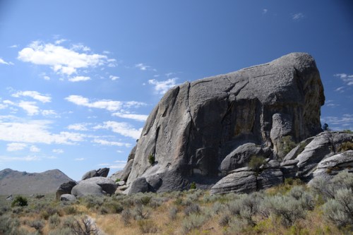 A large granite rock formation stands against a blue sky with scattered clouds, surrounded by sparse vegetation.