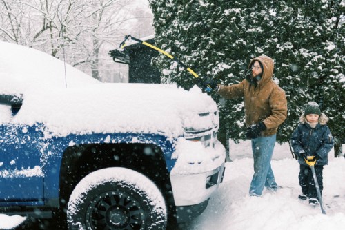 A man and a child clear snow from a blue truck in a snowy neighborhood. Trees are covered in snow in the background.