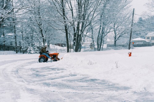 A person using a tractor to clear snow from a driveway in a snowy landscape with trees and houses in the background.