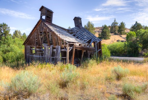 An old, dilapidated wooden building with a collapsed roof, surrounded by tall grass and trees under a clear blue sky.