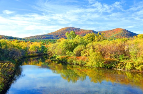 Scenic view of a river reflecting colorful autumn foliage and mountains under a blue sky with wispy clouds.