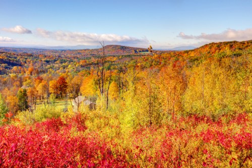 Vibrant autumn landscape with colorful trees in shades of red, orange, and yellow, overlooking a valley and distant hills.
