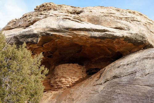 A rocky cliff with a large overhang and a circular stone structure nestled beneath it, surrounded by sparse vegetation.
