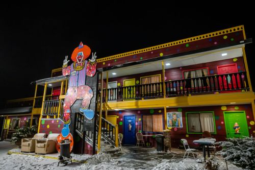 Colorful motel exterior featuring a large clown figure, snow on the ground, and brightly painted doors at night.