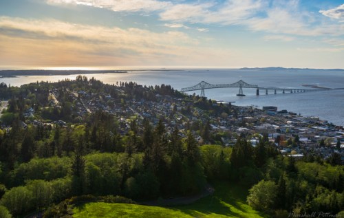Aerial view of a coastal town with a bridge, surrounded by greenery and water under a blue sky with clouds.