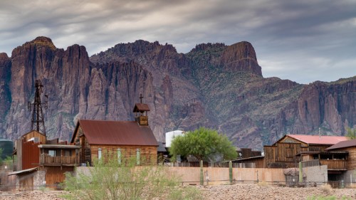 A rustic town with wooden buildings and a church, set against dramatic mountains under a cloudy sky.