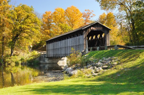 A wooden covered bridge spans a calm river, surrounded by vibrant autumn foliage and a grassy bank.