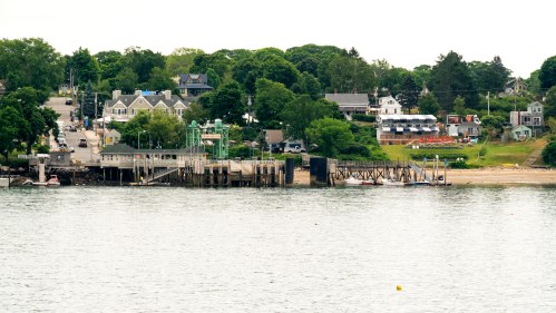 A coastal view featuring a pier, boats, and a tree-lined shore with houses in the background.