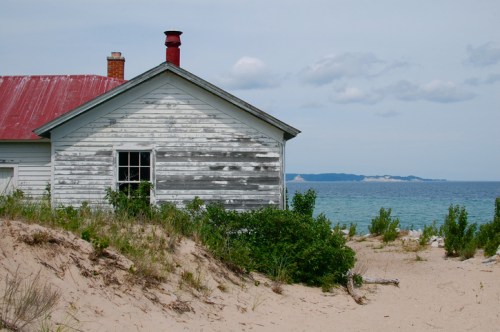 A weathered house with a red roof sits near a sandy beach, overlooking a calm lake and distant shoreline.
