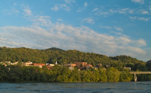 A scenic view of a small town nestled by a river, surrounded by green hills and a blue sky with scattered clouds.