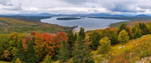 A panoramic view of a lake surrounded by vibrant autumn foliage and distant mountains under a cloudy sky.