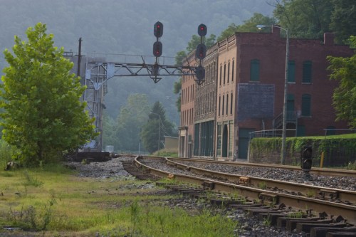 A railway track curves past a brick building, with signal lights overhead and greenery nearby, set against a misty backdrop.