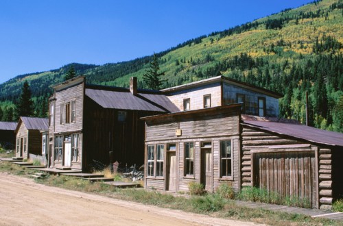 Abandoned wooden buildings line a dirt road, set against a backdrop of green mountains and clear blue sky.