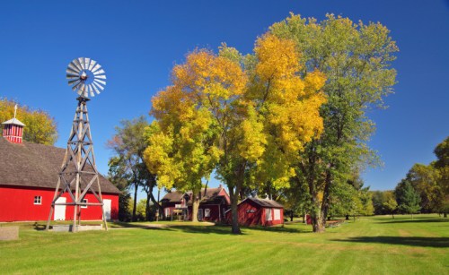 A red barn and small buildings surrounded by colorful autumn trees and a windmill under a clear blue sky.