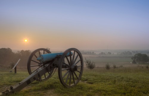 A historic cannon sits on a grassy hill at sunrise, overlooking a misty landscape.