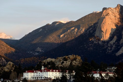 A grand white hotel nestled at the base of towering mountains, with rocky formations and trees surrounding the area.