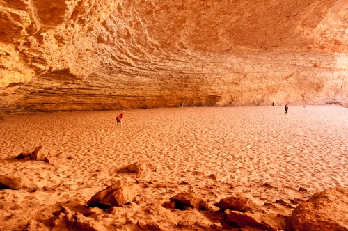 A sandy cave interior with textured rock walls and a few people exploring the area. Warm, earthy tones dominate the scene.