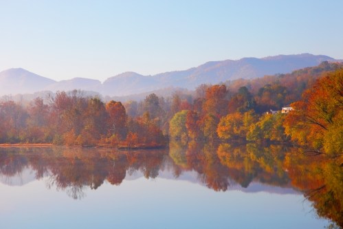 A serene lake reflects colorful autumn trees and distant mountains under a clear blue sky.