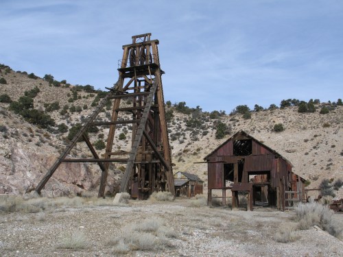 Abandoned mining structure with a wooden headframe and dilapidated building against a rocky hillside.