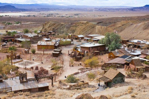 Aerial view of a rustic ghost town with wooden buildings, dirt paths, and surrounding desert landscape.
