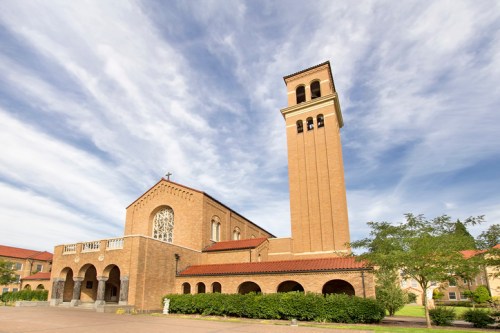 A brick church with a tall bell tower under a blue sky with clouds, surrounded by greenery.