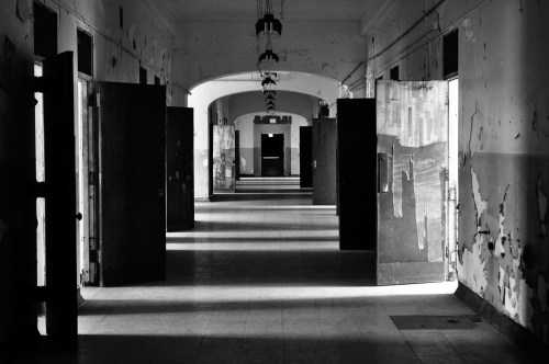 A dimly lit corridor with multiple open doors, featuring peeling paint and shadows creating a haunting atmosphere.