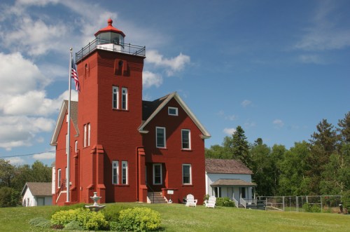 A red lighthouse with a white lantern room, surrounded by green grass and trees, under a blue sky with clouds.