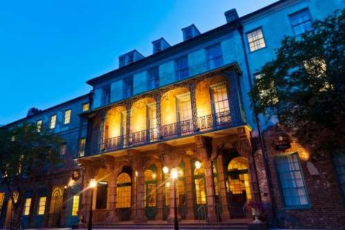Historic building with ornate balconies, illuminated at dusk, surrounded by trees and street lamps.