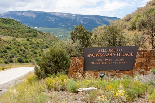 Sign welcoming visitors to Snowmass Village, surrounded by greenery and mountains under a partly cloudy sky.
