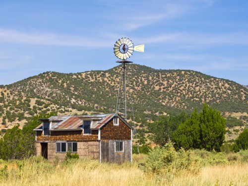 A rustic wooden house with a windmill stands in a grassy field, surrounded by rolling hills and blue skies.
