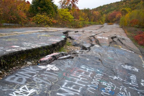 Cracked and damaged road covered in graffiti, surrounded by autumn foliage and trees.