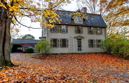 A charming two-story house surrounded by vibrant autumn leaves and trees, with a gravel driveway and a garage in the background.