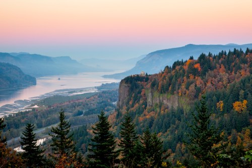 A serene landscape featuring a river valley, autumn foliage, and misty mountains at sunset.