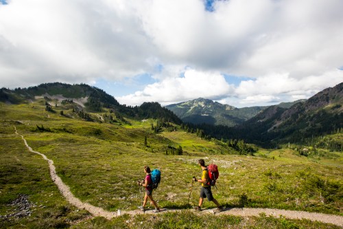 Two hikers with backpacks walk along a winding trail through a lush green landscape under a partly cloudy sky.